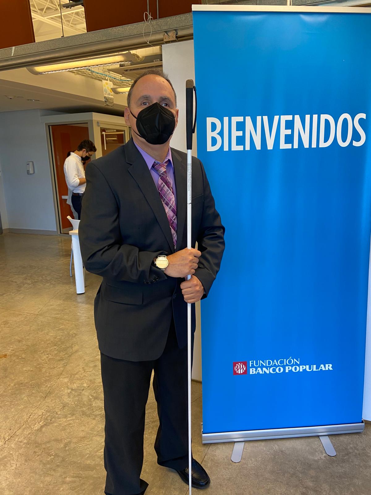 La foto presenta a Manolo posando al lado del letrero de la Fundación del Banco Popular.