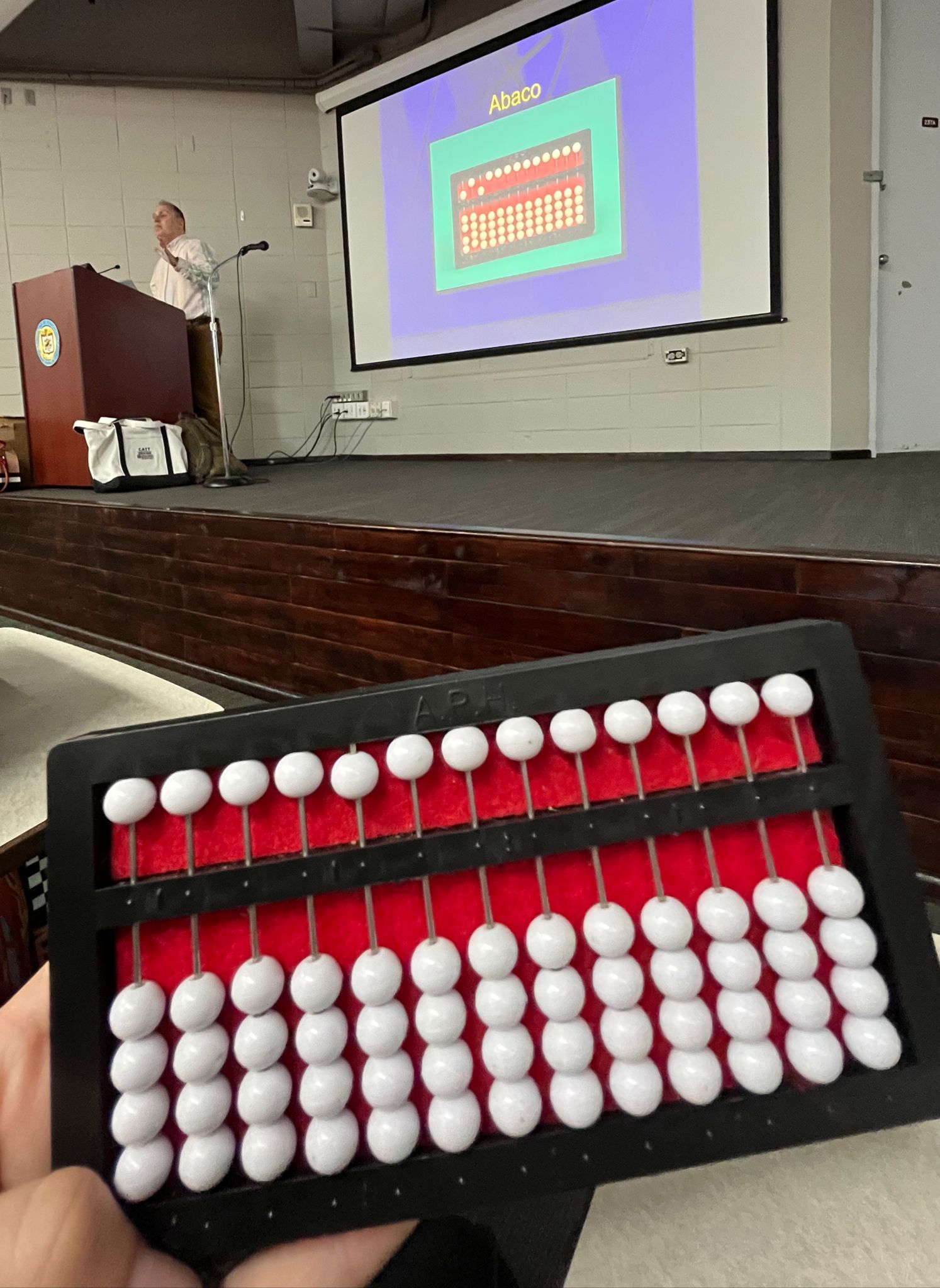 La foto presenta a Manolo en el podio de una tarima ofreciendo la charla relacionada a las matemáticas y los estudiantes ciegos.