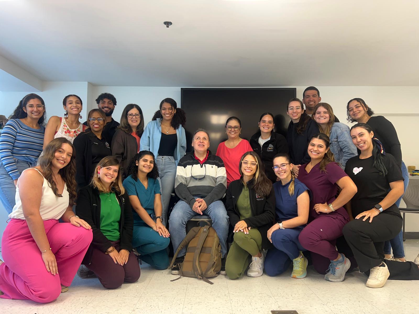 La foto presenta a Manolo sentado en el centro de un grupo de estudiantes de terapia ocupacional. Hay estudiantes que están de pie y otros de rodillas posando para la foto.