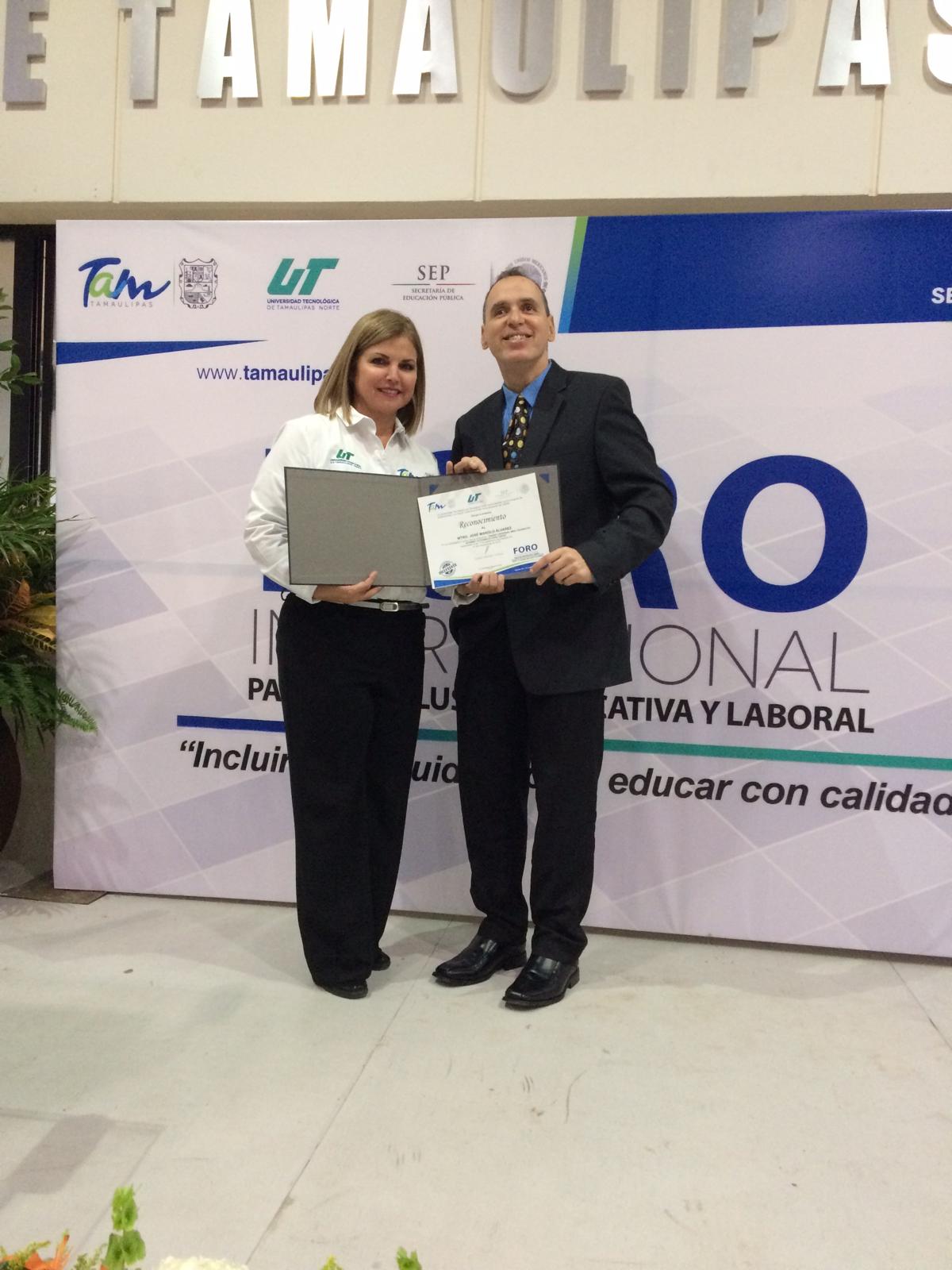 La foto muestra a Manolo recibiendo un certificado de una mujer en la tarima. La mujer a la izquierda lleva una camisa blanca con logotipos y sostiene un diploma o certificado. Mientas que Manolo está a la derecha y viste un traje oscuro con una corbata y también sostiene el diploma.