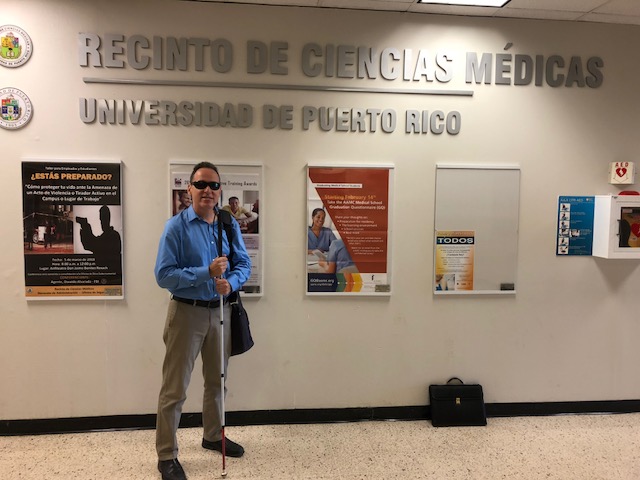 La foto presenta a Manolo posando al lado del letrero del Recinto de Ciencias Médicas de la UPR.